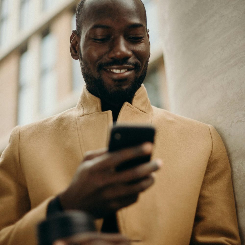 A cheerful man in a stylish coat using his smartphone and holding coffee outside.